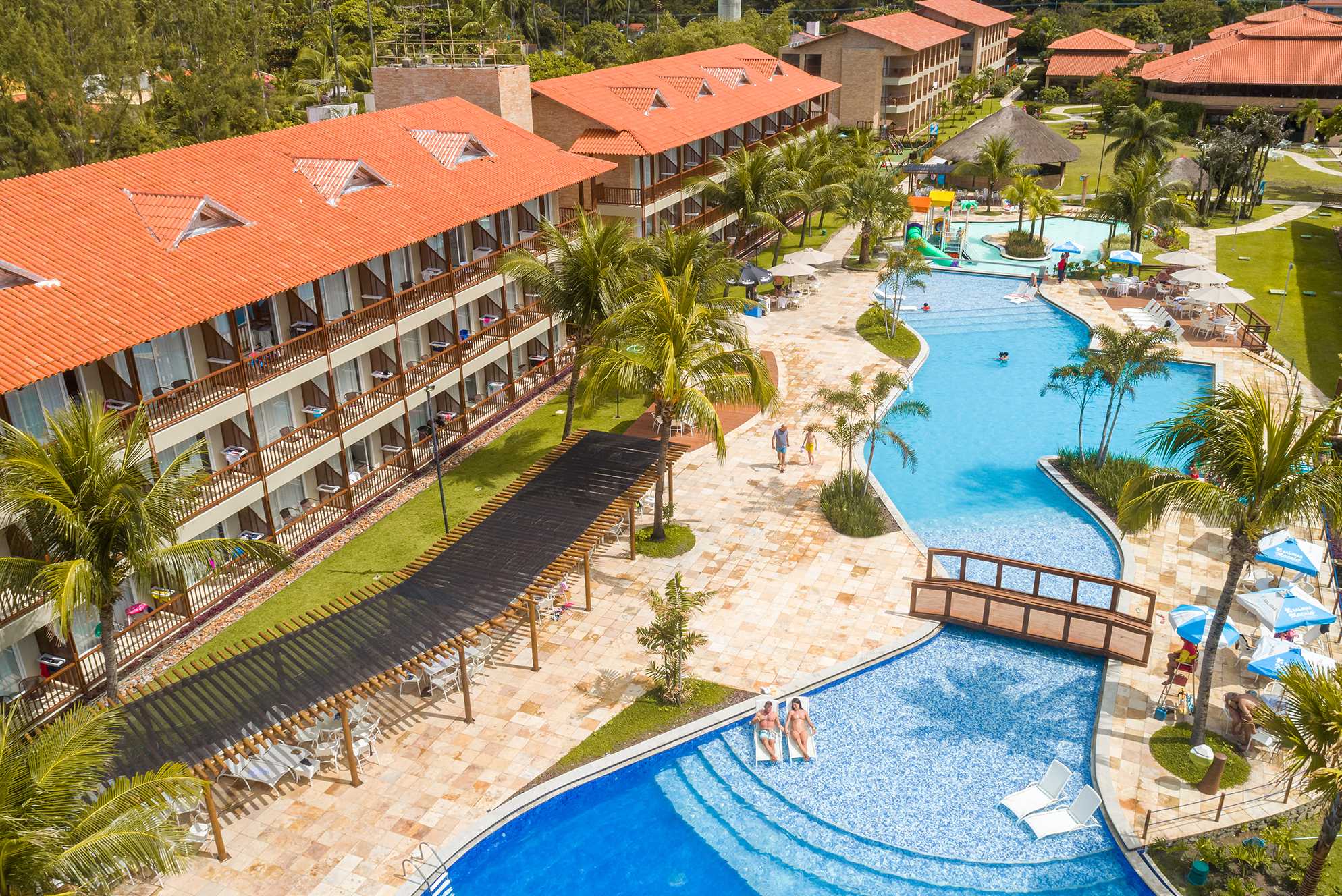 A imagem captura o Salinas Maceió, exibindo uma vista aérea das instalações do resort. Edifícios com telhados de terracota se alinham à beira da piscina, que é o ponto focal da área de lazer, rodeada por palmeiras e espreguiçadeiras.
