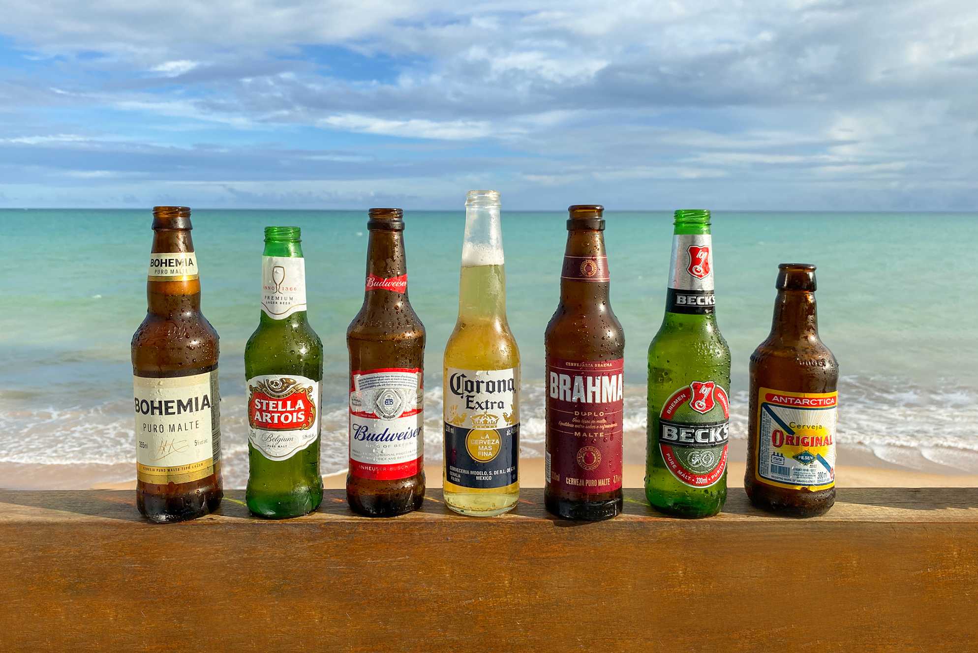 Frente ao horizonte azul do mar, garrafas de cervejas conhecidas — Bohemia, Stella Artois, Corona, Brahma, Beck's e Antárctica Original — estão molhadas por gotas de condensação.