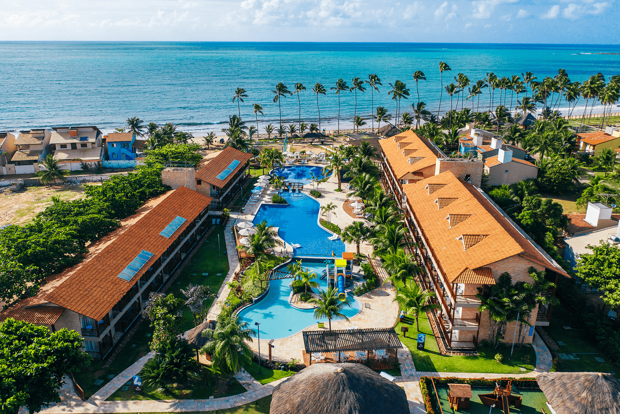 Vista aérea do resort Salinas Maceió. Cercado por palmeiras altas e diretamente de frente para a praia, com o vasto oceano azul ao fundo. As construções do resort têm telhados de cerâmica vermelha, e uma grande piscina em forma de lagoa com toboágua.
