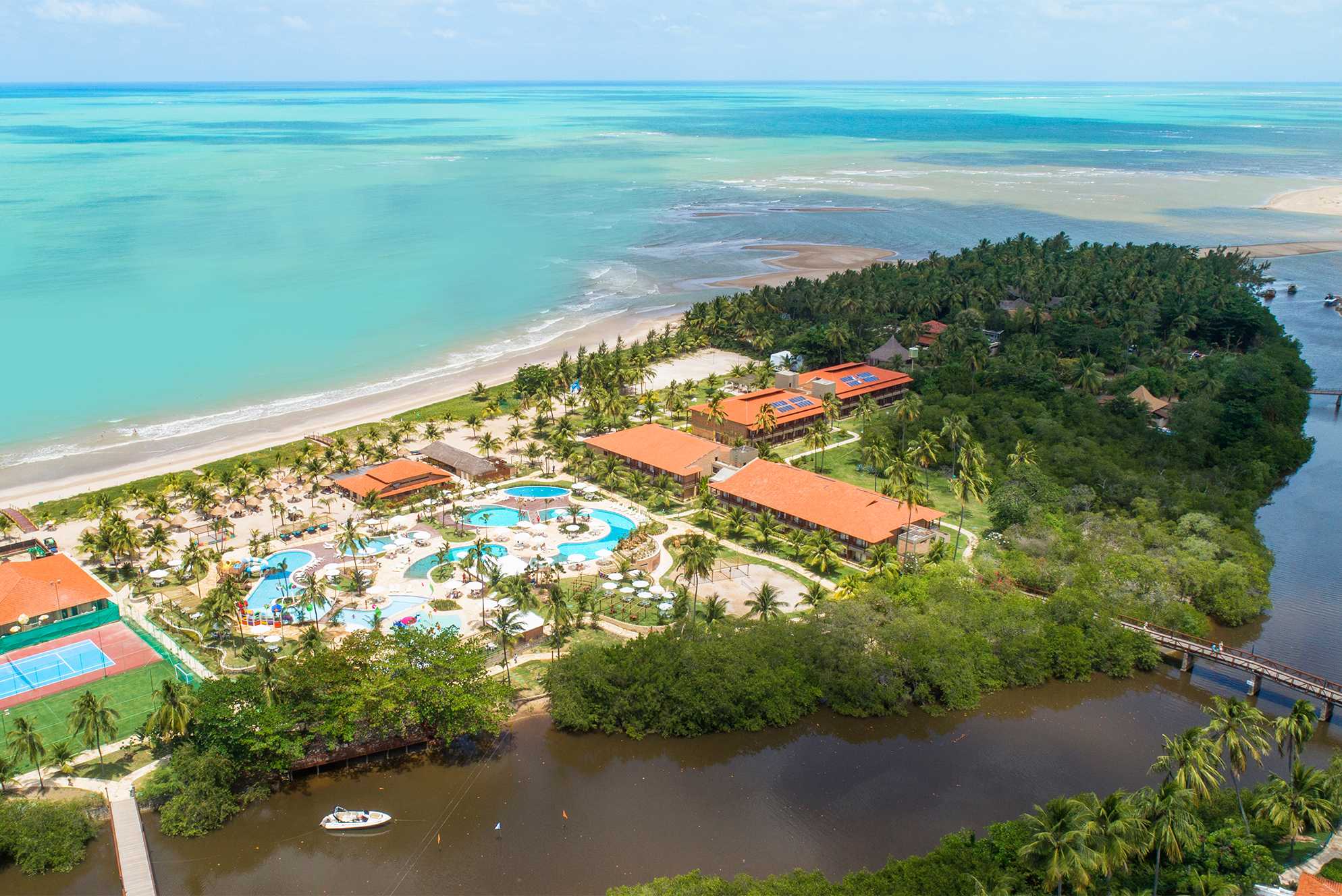 Vista aérea de um resort situado entre um rio e a praia de maragogi. O mar tem tons de azul e areia é branca. Há várias piscinas e edifícios no resort, que é cercado por vegetação tropical.