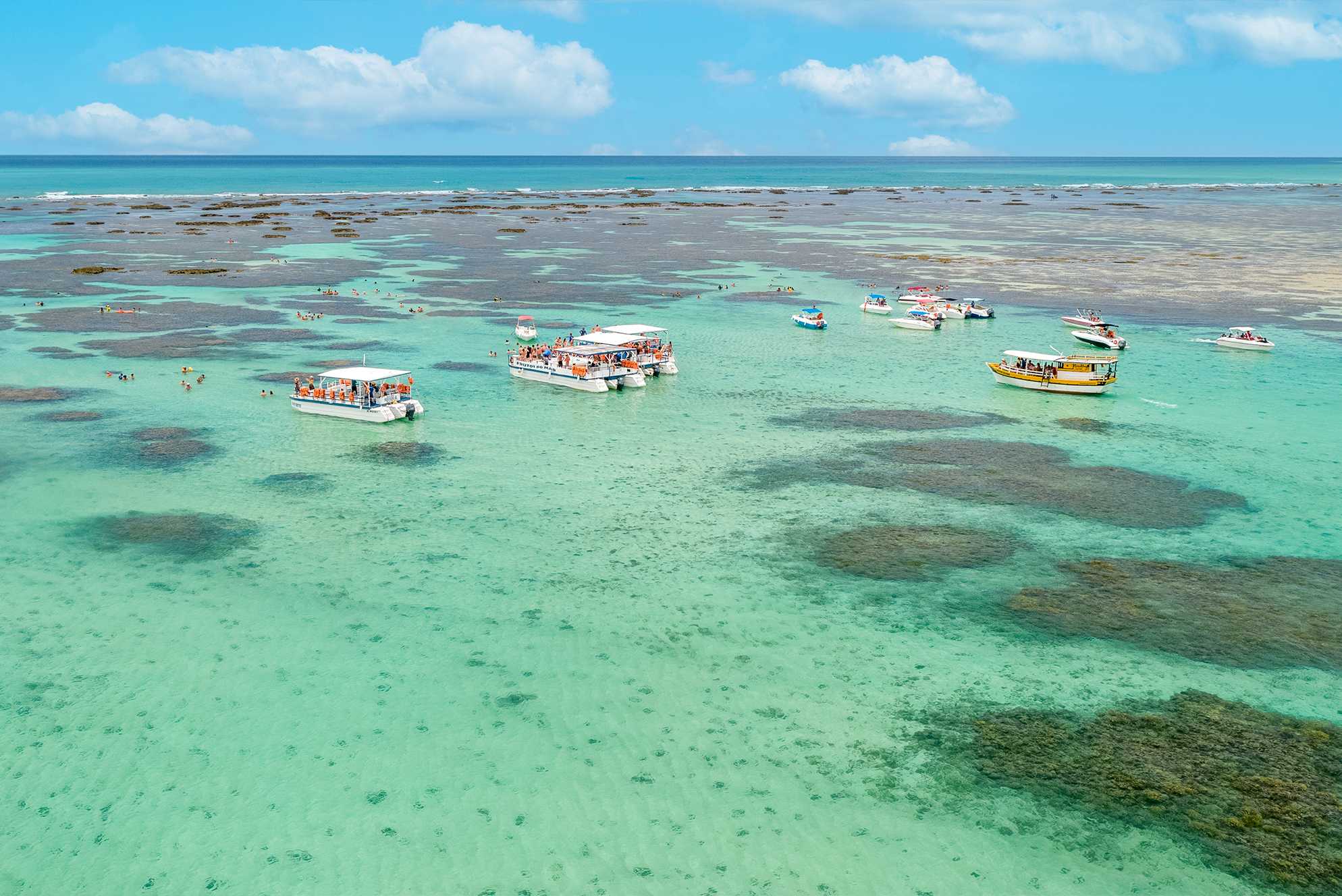 Vista aérea das piscinas naturais de Maragogi. Barcos estão ancorados nas águas, que variam em tons de verde claro a azul. As formações de recifes são visíveis sob a superfície do mar. O céu é amplo e azul com algumas nuvens brancas.