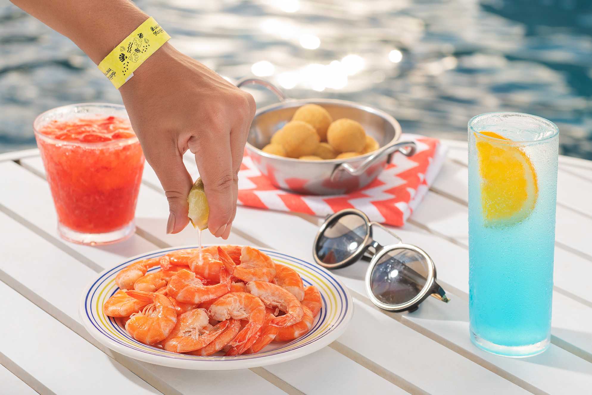 A foto captura um momento culinário ao ar livre, com uma mão feminina espremendo uma fatia de limão sobre uma porção de camarões em um prato decorado. Há também uma panela de coxinhas e um guardanapo xadrez. Óculos de sol repousam ao lado.