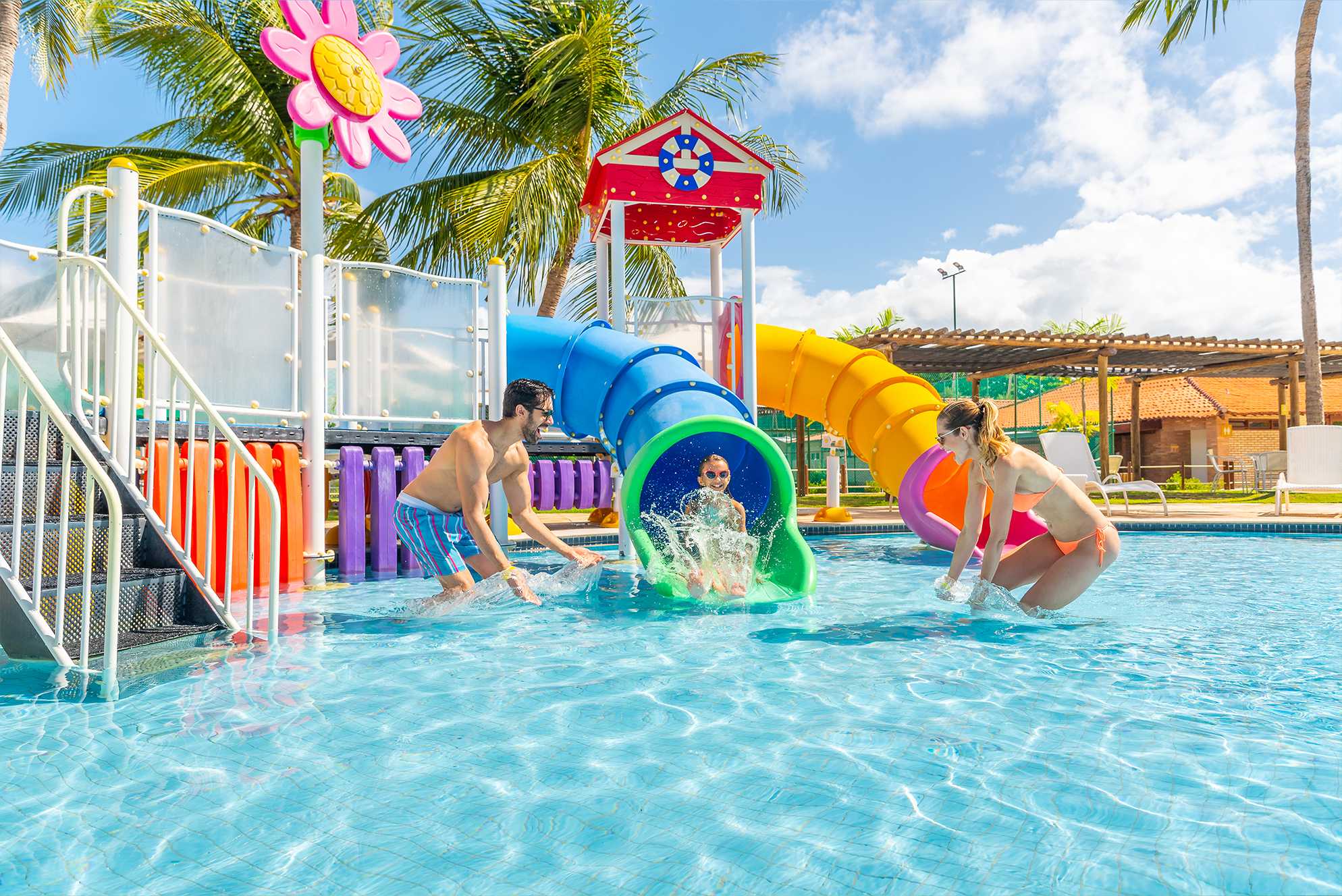 Em uma piscina infantil uma criança sorridente desce por um escorregador aquático azul, fazendo um grande splash ao entrar na água, onde um homem e uma mulher estão agachados esperando por ela. Ele é cercado por estruturas de playground coloridas.