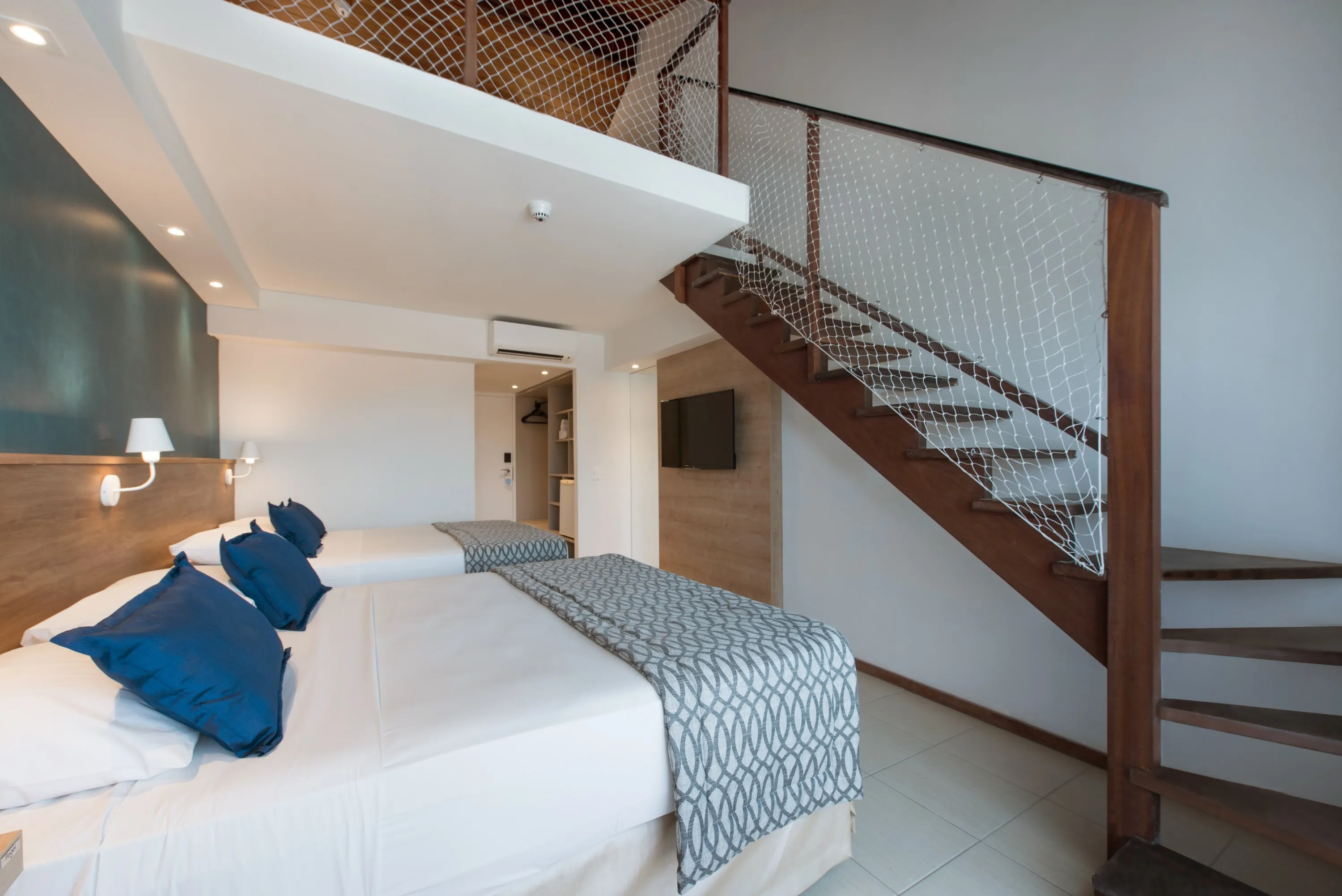 Planta baja de la acomodación Suite Familiar, en la versión con mezanino. Dos camas queen size, dos luminarias, siendo una para cada cama, painel de madera con TV por cable,  espejo, ropero y escalera llevándonos al primer piso.