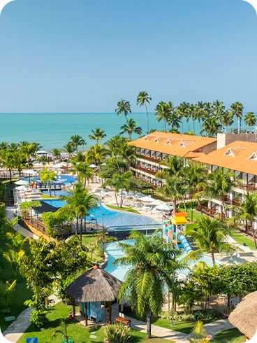 Vista panorámica del resort Salinas Maceió, que muestra los edificios de apartamentos, el complejo de piscinas rodeado de cocoteros y vegetación, y el mar turquesa al fondo en un día soleado.