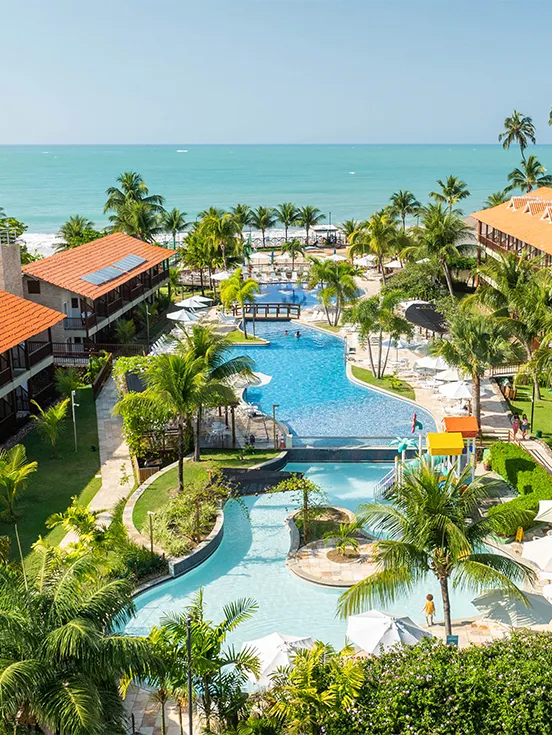 Vista panorámica del resort Salinas Maceió, que muestra los edificios de apartamentos, el complejo de piscinas rodeado de cocoteros y vegetación, y el mar turquesa al fondo en un día soleado.