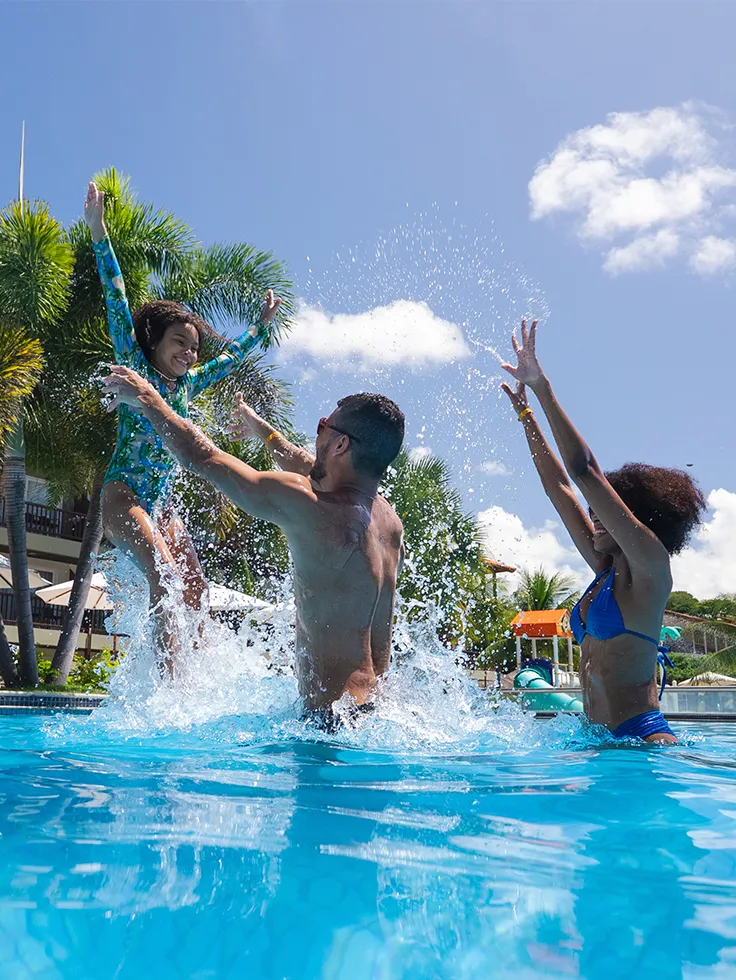 Criança brincando com seus pais na piscina infantil, o pai está jogando ela pro alto. Ao fundo tem a estrutura do resort.
