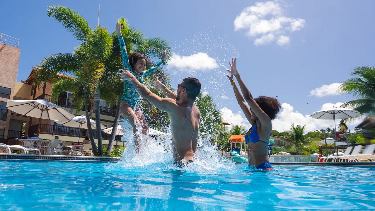 Criança brincando com seus pais na piscina infantil, o pai está jogando ela pro alto. Ao fundo tem a estrutura do resort.