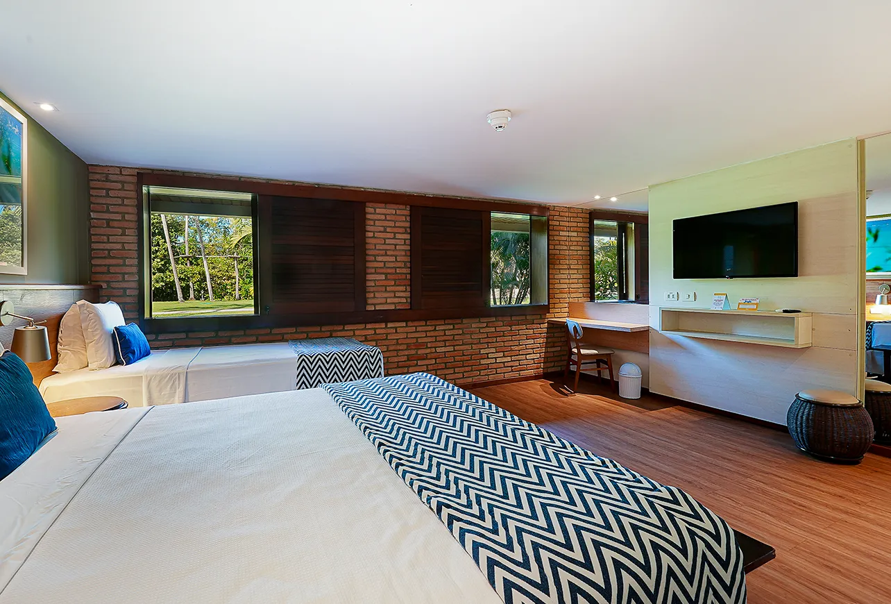 Uma cama queen size e uma cama solteiro na Suíte Familiar. Há um grande espaço vazio entre as camas e a parede com escrivaninha, painel com televisão e espelho. Tem janelas abertas pro jardim do Salinas Maragogi.