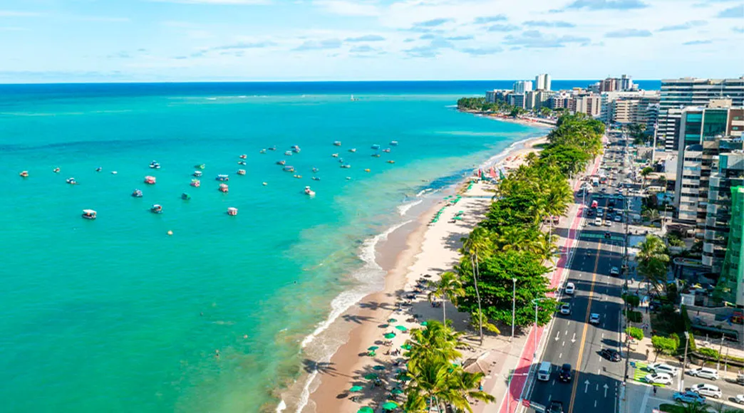 Vista aérea da orla de Maceió na Praia de Jatiúca, mostrando o mar verde com muitos barcos de passeio, a faixa de areia com coqueiros, a avenida costeira movimentada e os altos prédios da cidade.
