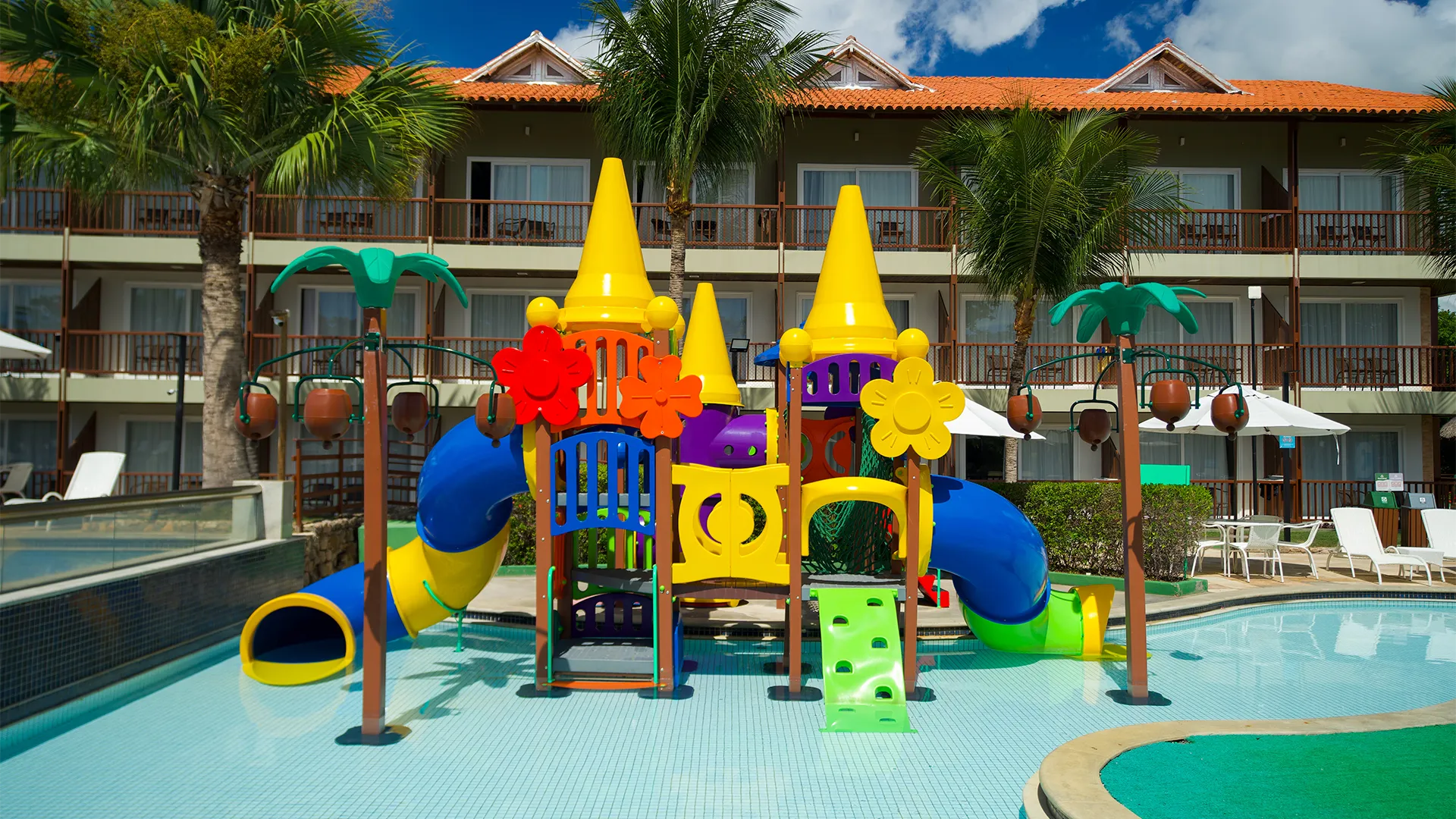 Brinquedão infantil na piscina infantil do Salinas Maceió. Ao fundo os apartamentos do resort com varandas, alguns coqueiros e área de lazer.