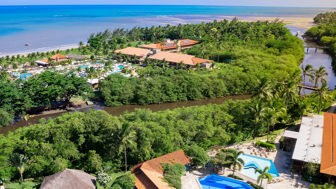 Panoramic aerial view of the Salinas Maragogi resort, showcasing its privileged location between the sea and the Maragogi River. The image showcases the hotel buildings, the water park, the bridges crossing the river, and the dense surrounding vegetation.