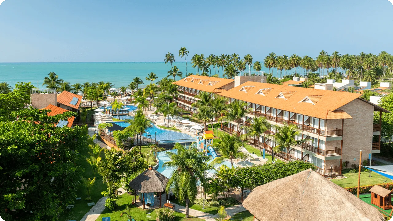 Vista panorámica del resort Salinas Maceió, que muestra los edificios de apartamentos, el complejo de piscinas rodeado de cocoteros y vegetación, y el mar turquesa al fondo en un día soleado.