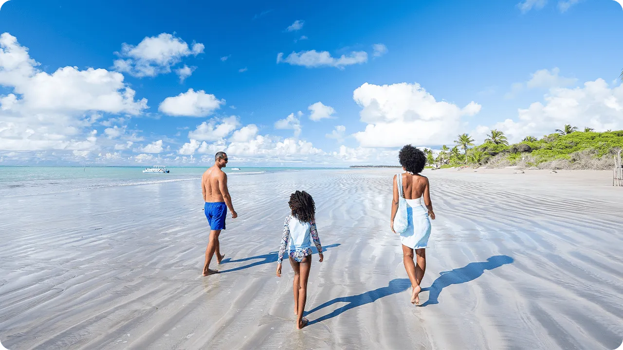Família curtindo dia ensolarado na praia de Ipioca andando descalços na areia clara, com mar azul-turquesa e uma pequena vegetação verde ao lado.