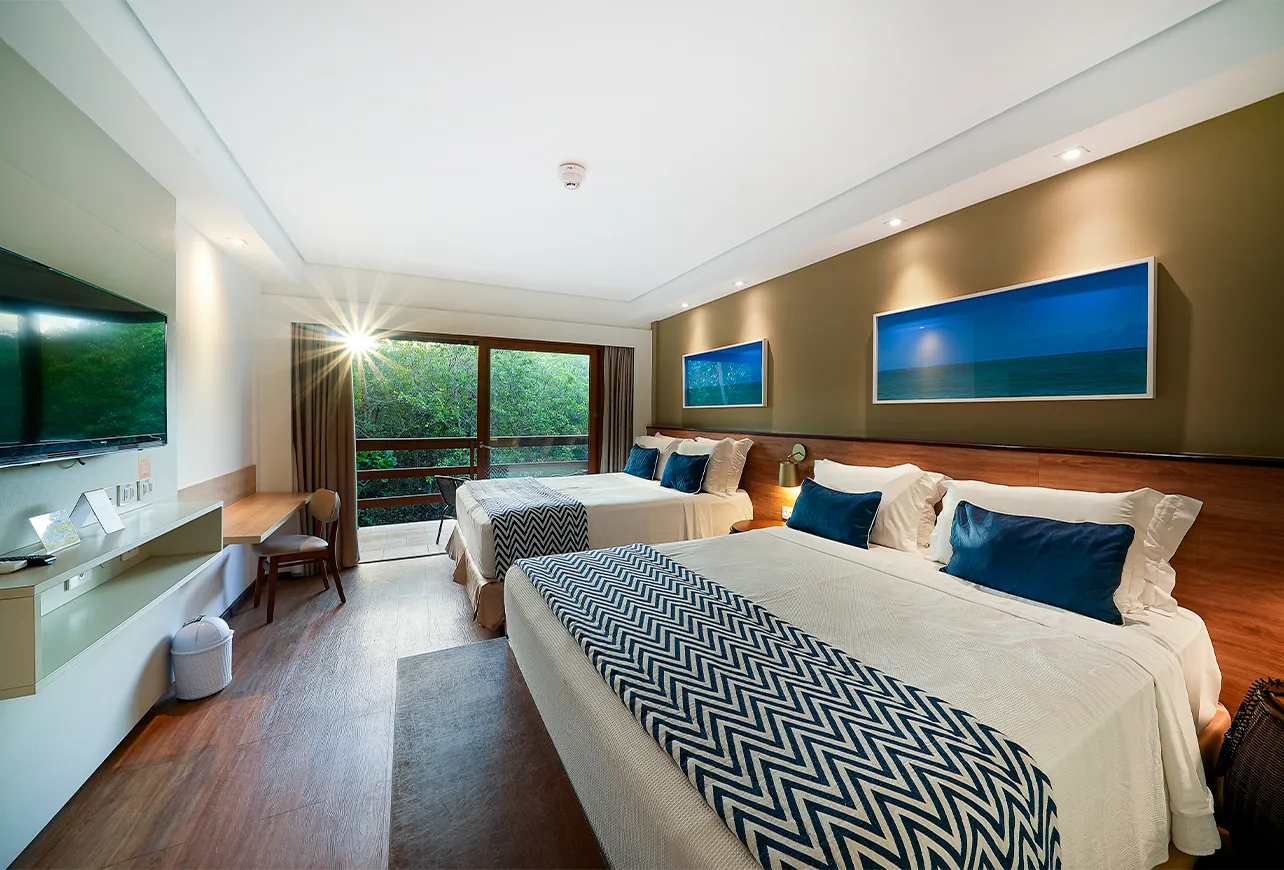 Apartamento Praia Jardim, contendo duas camas queen size, quadros pendurados na parede de tom verde. Na frente das camas, tem uma televisão pendurada, uma escrivaninha e, ao fundo, a varanda com vista pro jardim.