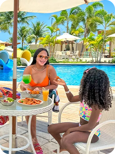 Três pessoas sentadas à mesa redonda sob ombrelone, com petiscos e bebidas; piscina e toboáguas azuis ao fundo, palmeiras e céu claro. Um homem segura uma cerveja, a mulher um drink verde e a criança prova um salgado.