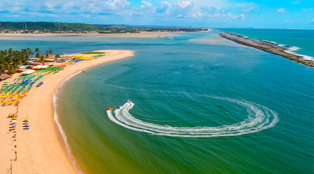 Vista aérea da Praia do Gunga, que é uma ponta de areia com coqueiros separando o mar aberto à direita e a Lagoa do Roteiro à esquerda. Um jet ski faz um círculo na água da lagoa.
