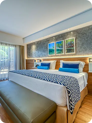 Interior del apartamento Estándar Vitória-Régia, con cama queen size, mesa y silla, decoración acogedora con detalles de madera, marcos decorativos con fotografías de las playas de Maragogi y balcón con vista al jardín.