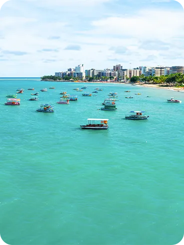 Barcos coloridos flutuando em águas turquesa na orla da praia de Jatiúca, com prédios à beira-mar e palmeiras na areia.