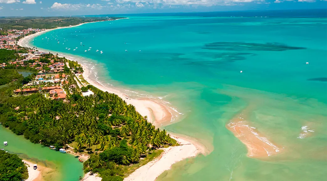 Vista aérea da Praia de Maragogi, com águas cristalinas e ao fundo a cidade e vegetação.