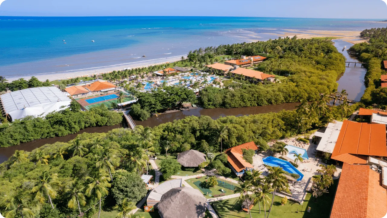 Vista aérea panorámica del resort Salinas Maragogi, que muestra su ubicación privilegiada entre el mar y el río Maragogi. La imagen muestra los edificios del hotel, el parque acuático, los puentes que cruzan el río y la densa vegetación circundante.