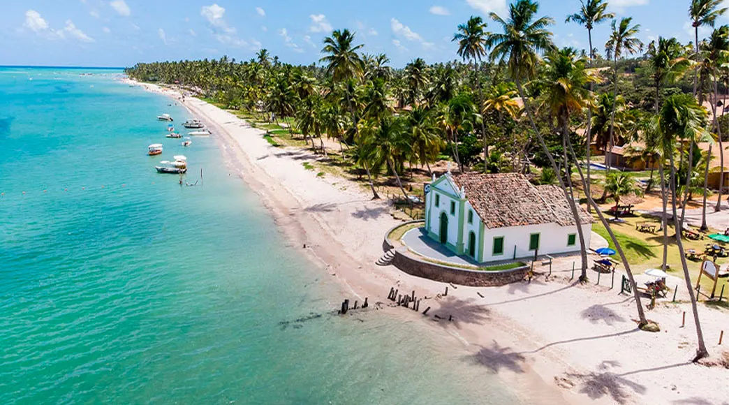 Vista aérea da Capela de São Benedito, uma igrejinha branca localizada na areia da Praia dos Carneiros, cercada por um extenso coqueiral e pelo mar calmo de águas esverdeadas com barcos ancorados.