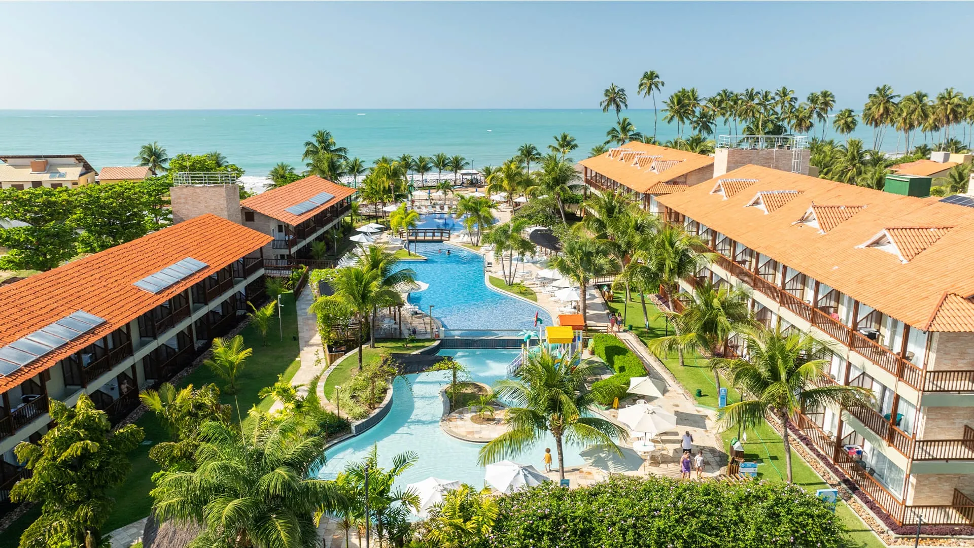 Vista panorámica del resort Salinas Maceió, que muestra los edificios de apartamentos, el complejo de piscinas rodeado de cocoteros y vegetación, y el mar turquesa al fondo en un día soleado.