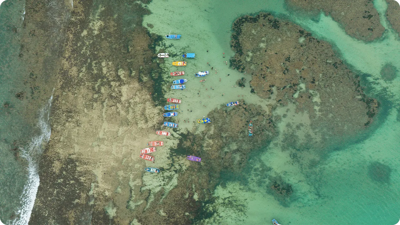 Barcos coloridos flutuando sobre águas rasas e cristalinas na Praia de Pajuçara, com recifes de corais visíveis, com maré baixa.