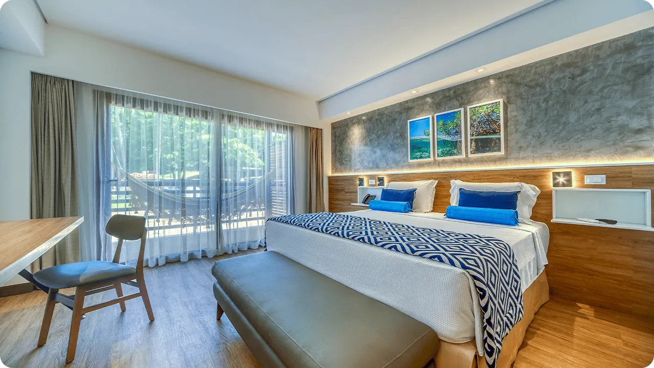 Interior del apartamento Estándar Vitória-Régia, con cama queen size, mesa y silla, decoración acogedora con detalles de madera, marcos decorativos con fotografías de las playas de Maragogi y balcón con vista al jardín.