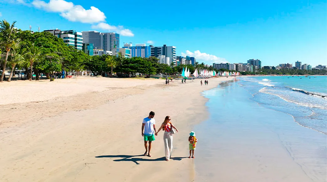 Família com uma criança caminha de mãos dadas pela areia molhada da Praia de Pajuçara. Ao fundo, a orla de Maceió é vista com seus prédios e as tradicionais jangadas na praia.