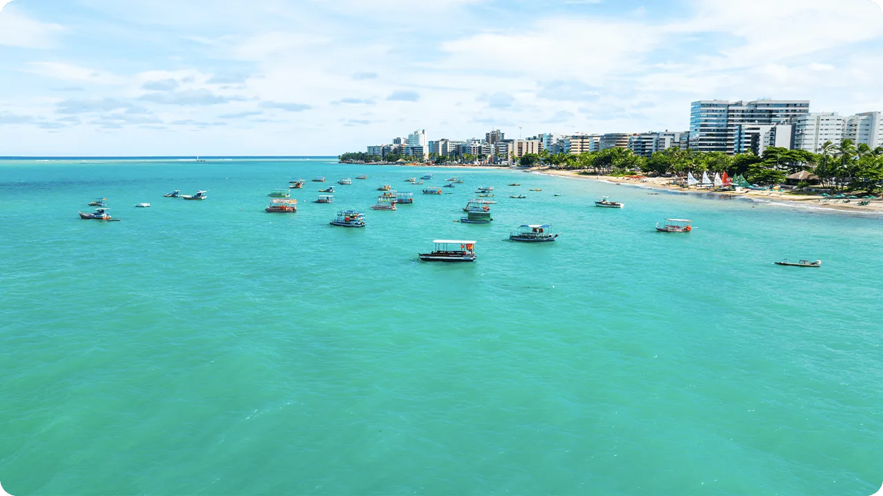 Barcos coloridos flutuando em águas turquesa na orla da praia de Jatiúca, com prédios à beira-mar e palmeiras na areia.