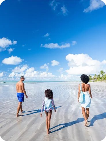 Família curtindo dia ensolarado na praia de Ipioca andando descalços na areia clara, com mar azul-turquesa e uma pequena vegetação verde ao lado.