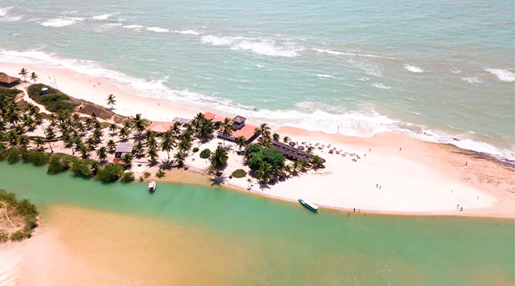 Vista aérea de Jequiá da Praia, mostrando uma faixa de areia com coqueiros que separa o rio de águas calmas e verdes do mar aberto com suas ondas.