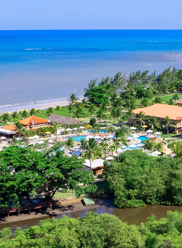 Panoramic aerial view of the Salinas Maragogi resort, showcasing its privileged location between the sea and the Maragogi River. The image showcases the hotel buildings, the water park, the bridges crossing the river, and the dense surrounding vegetation.