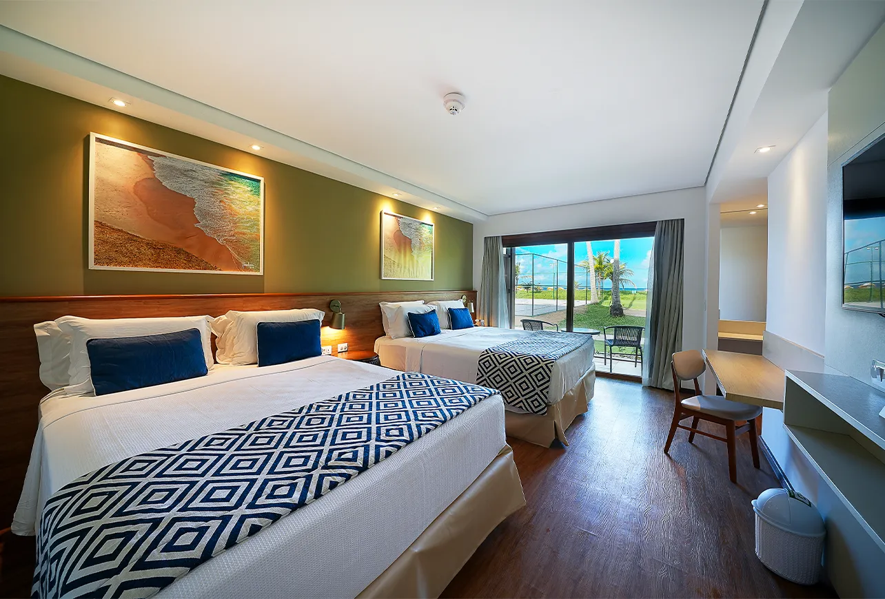 Detalhes da acomodação Praia Frente Mar, com duas camas queen size, mesa com cadeira, painel com televisão pendurada, quadros com fotografias de praias de Maragogi e uma varanda com vista pro mar e pro jardim.