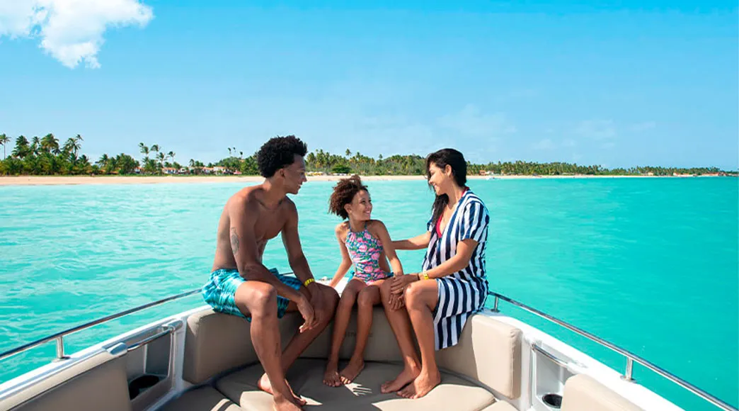 Família com pai, mãe e filha sentados na proa de uma lancha, conversando e sorrindo durante um passeio no mar de águas calmas e azul-turquesa, com a orla da Praia de Burgalhau e seus coqueiros ao fundo.