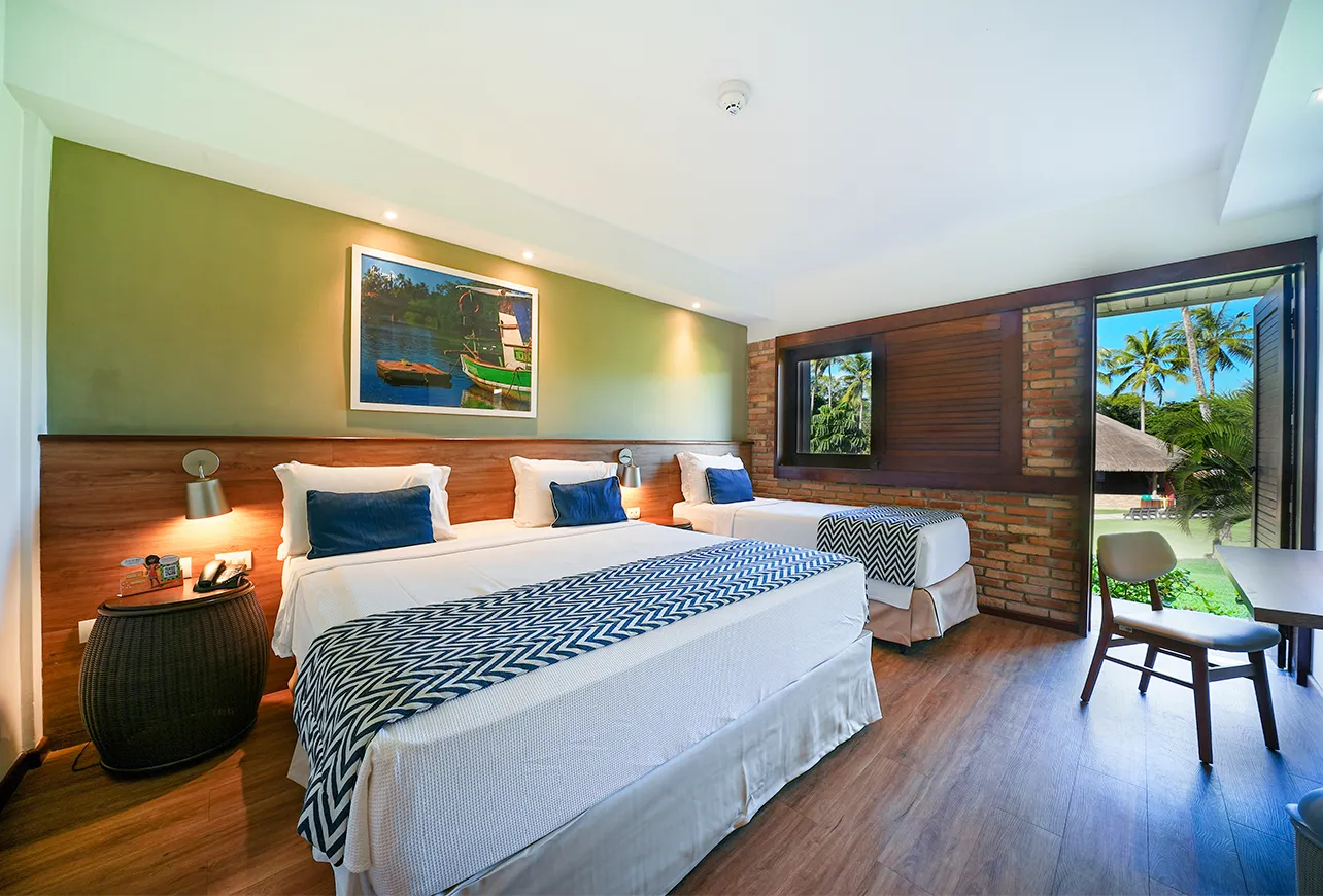 Apartamento Standard, uma das acomodações do Salinas Maragogi, com uma cama queen size e uma cama solteiro e decoração aconchegante com detalhes em madeira, luminárias e um quadro de praia de Maragogi. Ao fundo, a porta pra varanda com vista pro jardim.
