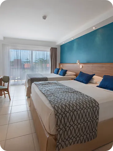 Two queen-size beds placed side by side, with two lamps, one for each bed, in front of a wooden panel with cable TV, a desk, and a chair.