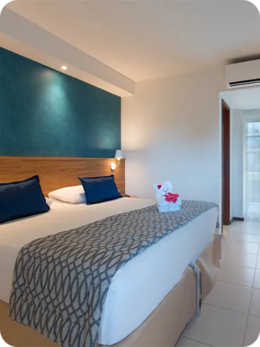 Cama queen size na acomodação Standard, decorada com ursinho  feito de toalha, em frente a roupeiro e painel de madeira com TV a cabo, mesa e cadeira.