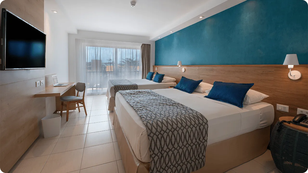 Two queen-size beds placed side by side, with two lamps, one for each bed, in front of a wooden panel with cable TV, a desk, and a chair.