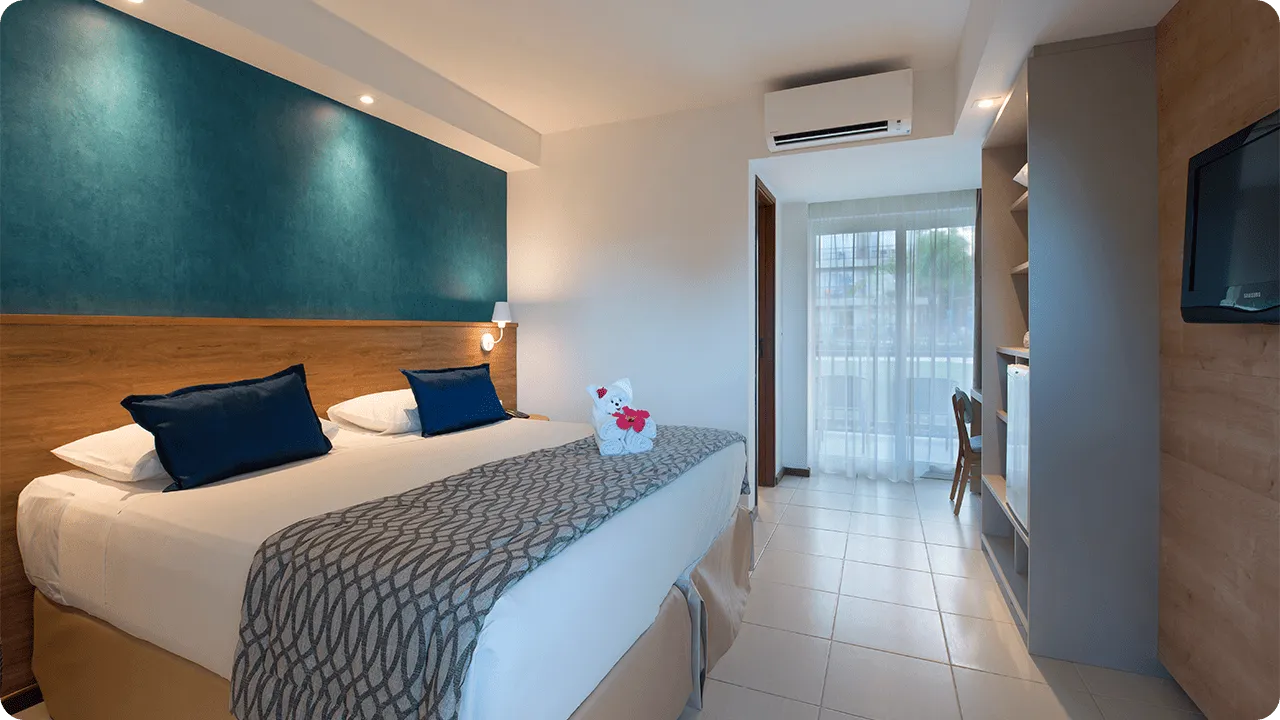 Cama queen size na acomodação Standard, decorada com ursinho  feito de toalha, em frente a roupeiro e painel de madeira com TV a cabo, mesa e cadeira.