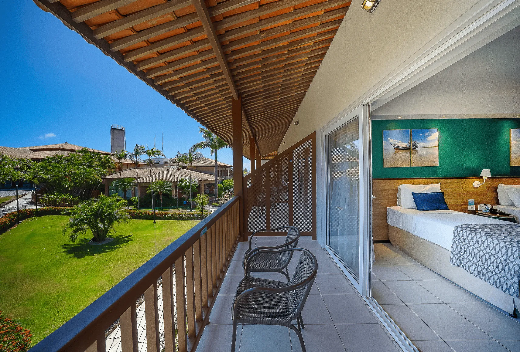 Varanda do Apartamento Standard no Salinas Maceió, ao lado esquerdo a vista pra jardins e área verde do resort e ao lado direito a parte interna do quarto com cama de solteiro.