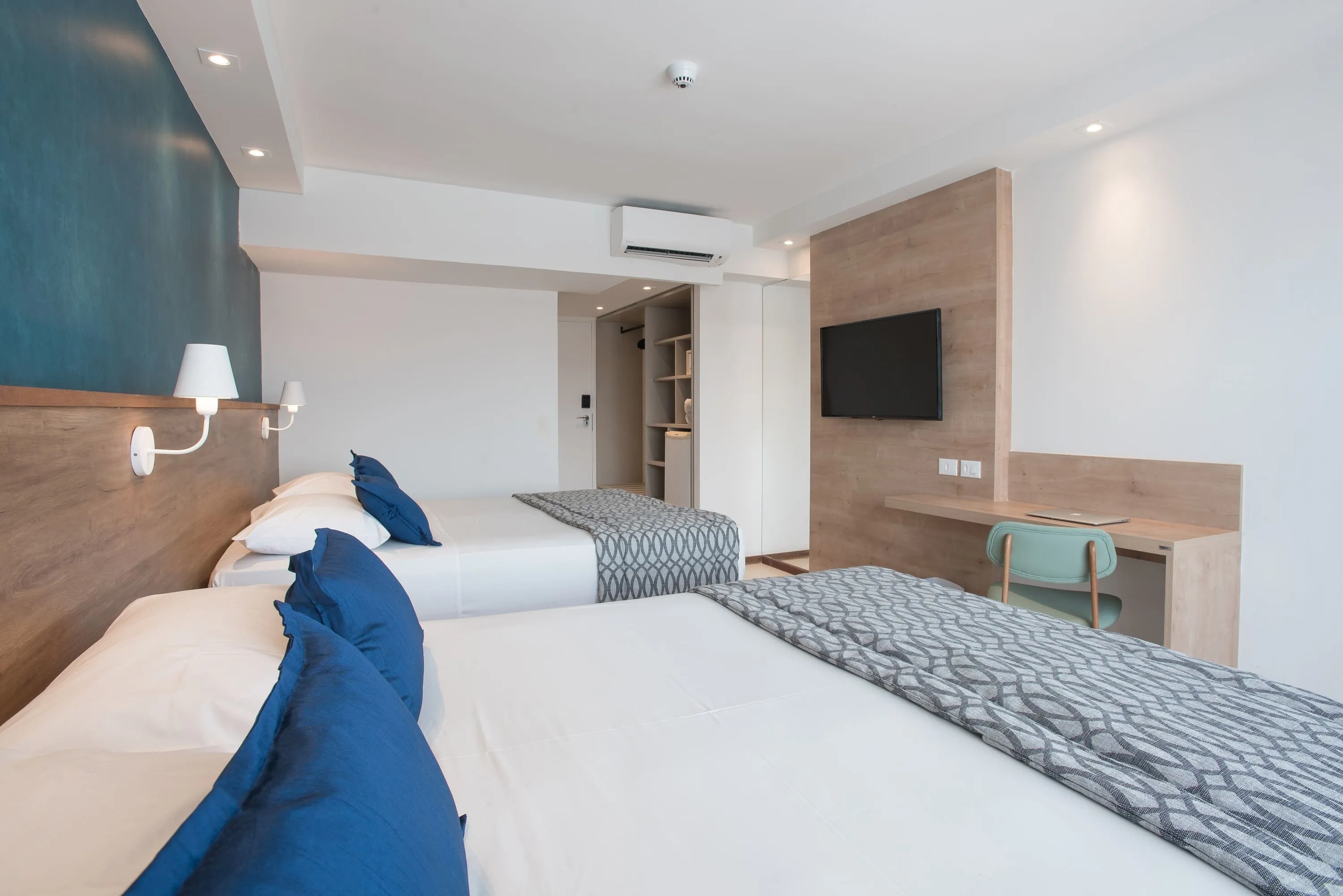 Puerta de entrada de la  acomodación Superior, ao lado del ropero y de dos camas queen size, dispuestas lado a lado, dos luminarias, siendo una para cada cama, en frente a painel de madera com TV por cable, mesa y silla.