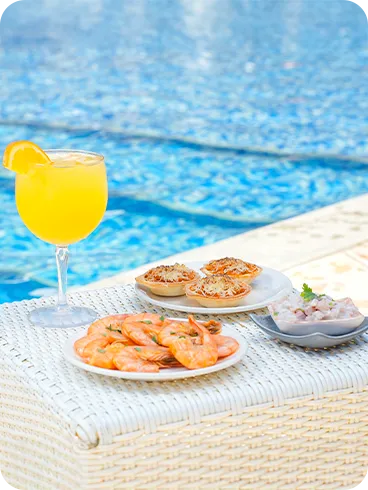 Mesa junto a la piscina con refresco de naranja y aperitivos, incluyendo camarones hervidos y mini tartas saladas, con el agua azul de la piscina de fondo.