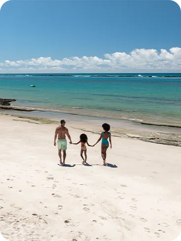 Família caminhando de mãos dadas na areia branca da Praia do Francês com mar e céu azul ao fundo e pequenas algas na faixa de areia.