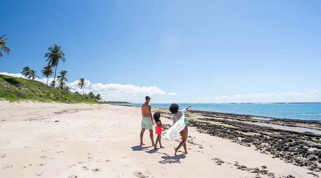 Família com pai, mãe e filho brincando nas ondas na beira do mar da Praia do Francês. A mãe esta de mãos dada com a filha e ela está apontando pro horizonte do mar e o pai acompanha em dia ensolarado.