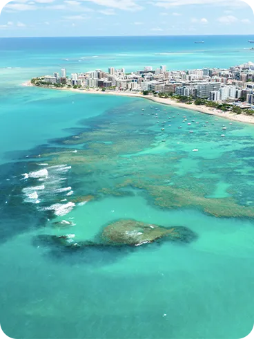 Vista aérea da Praia de Ponta Verde com mar azul cristalino, recifes de corais, pequenos barcos e prédios à beira-mar