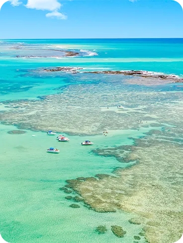 Vista aérea da Praia de Barra Grande com maré baixa e cristalina, recifes de corais visíveis sob a superfície, céu com pequenas nuvens do lado esquerdo e barcos espalhados na água.