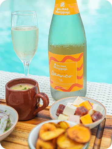 Garrafa de espumante do Salinas Maragogi e taça cheia sobre mesa à beira da piscina, com petiscos variados em tigelas sobre uma tábua.