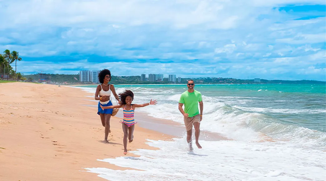 Família feliz com pai, mãe e filha correndo na beira do mar na Praia de Jatiúca, espirrando água das ondas. Ao fundo, a silhueta dos prédios da cidade é visível.